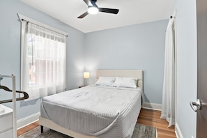 Une chambre à coucher avec un lit, un ventilateur de plafond, une fenêtre avec des rideaux et une table de chevet avec une lampe.