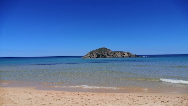 Ein Strand mit klarem Wasser, einem Sandufer und einem Felsen im Hintergrund.