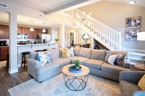 A modern living room with a sectional sofa, a coffee table, and a staircase leading to an upper level. - Spacious 3 BR 2 Bath Loft Condo in Grand Landing (Grand Haven)