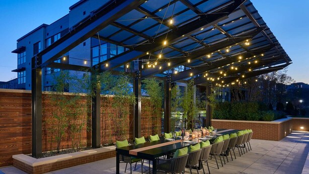 An outdoor patio with a long table and chairs, a glass ceiling structure, and a brick wall with greenery. - Global Luxury Suites Clarendon Metro (Arlington)