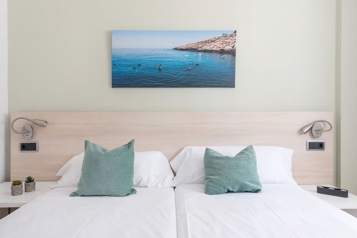A hotel room with a bed, two pillows, a wall-mounted picture of a beach, and bedside lamps.
