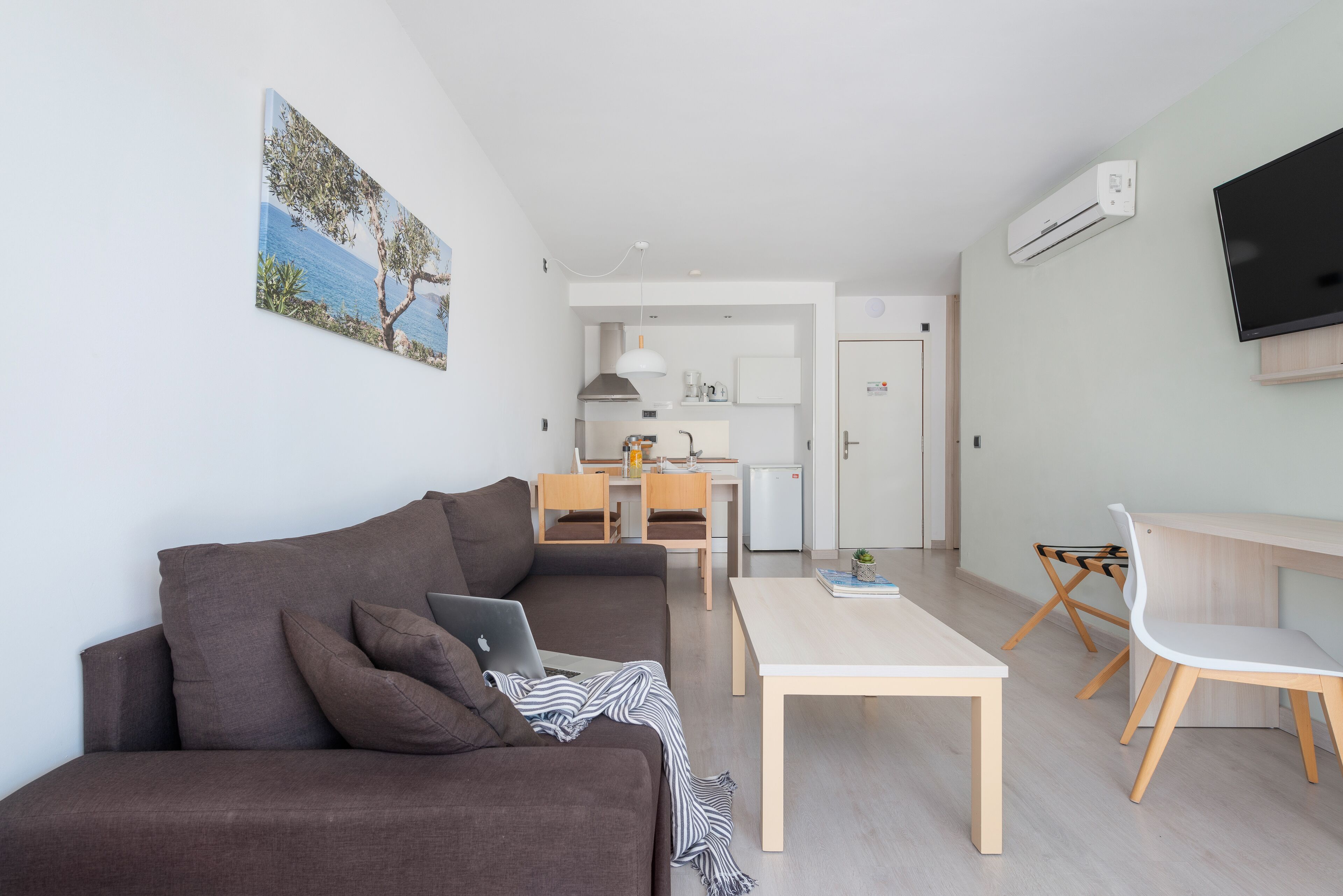 Un salon moderne avec un canapé, une table basse et un téléviseur. On aperçoit, au fond, un coin cuisine avec un réfrigérateur et un four à micro-ondes.