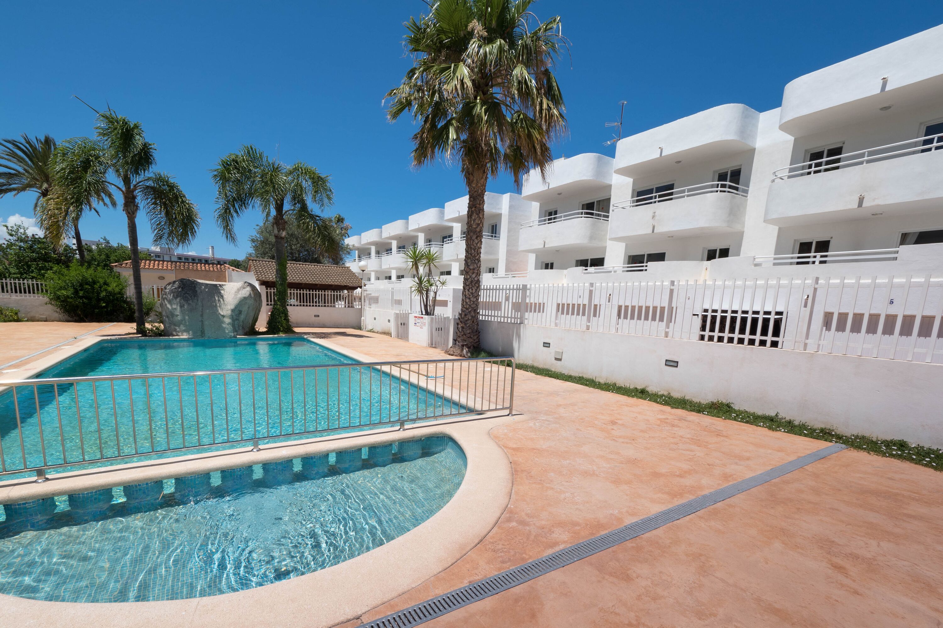 Una piscina de borde curvo, rodeada por una valla blanca, palmeras y un edificio con balcones.