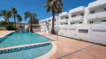 Una piscina de borde curvo, rodeada por una valla blanca, palmeras y un edificio con balcones.