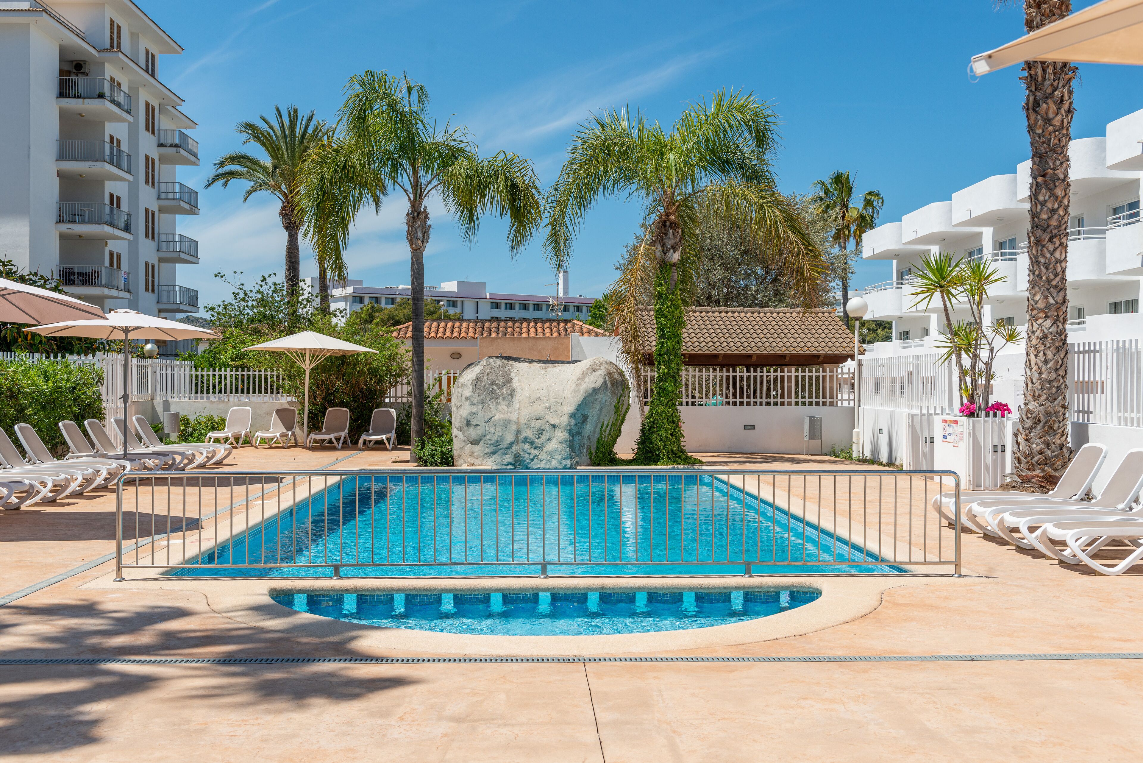 Una piscina con una formación rocosa en el centro, rodeada de palmeras y sillas de descanso.