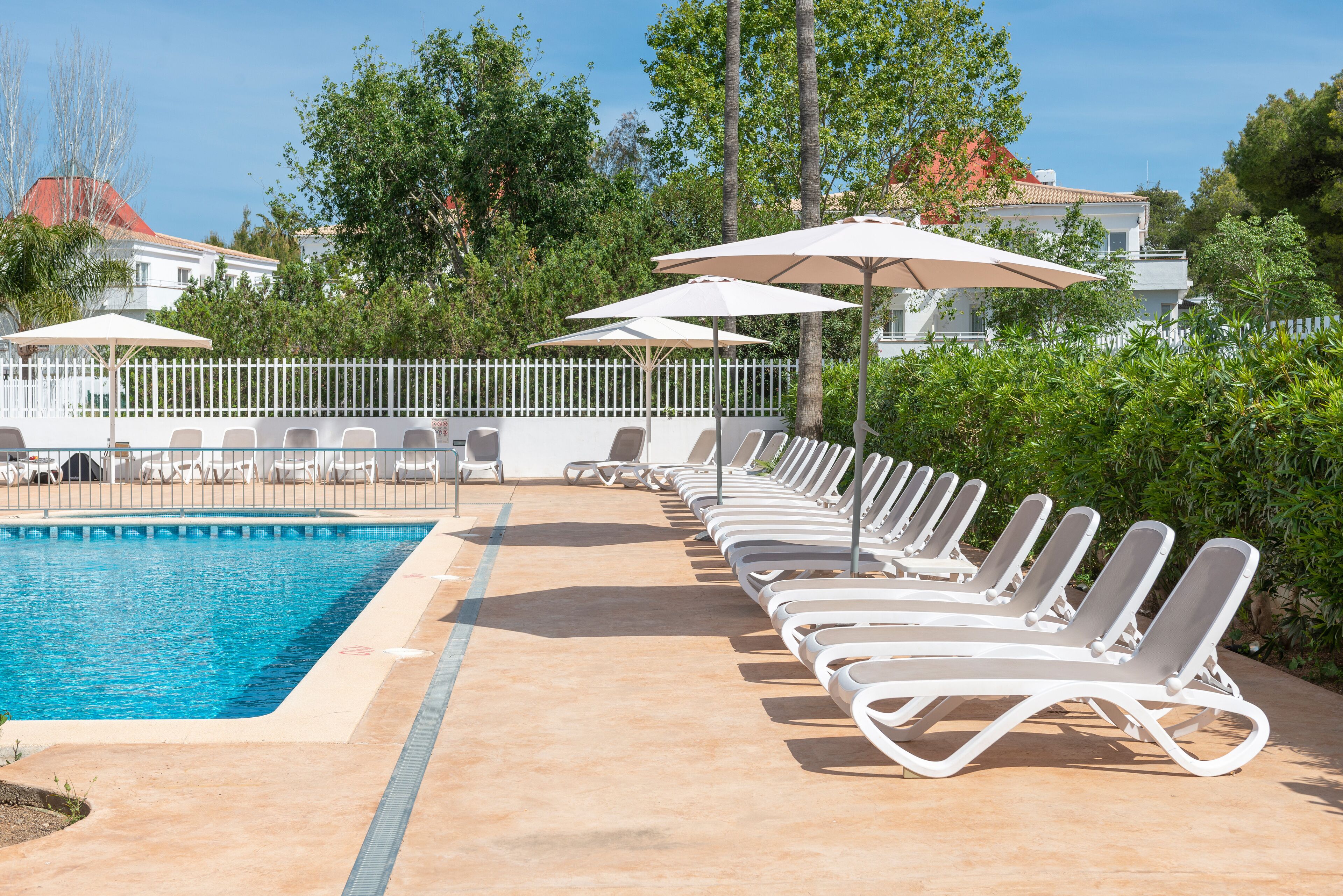 Zona de piscina con sillones de playa y sombrillas, rodeada por una valla blanca y vegetación.