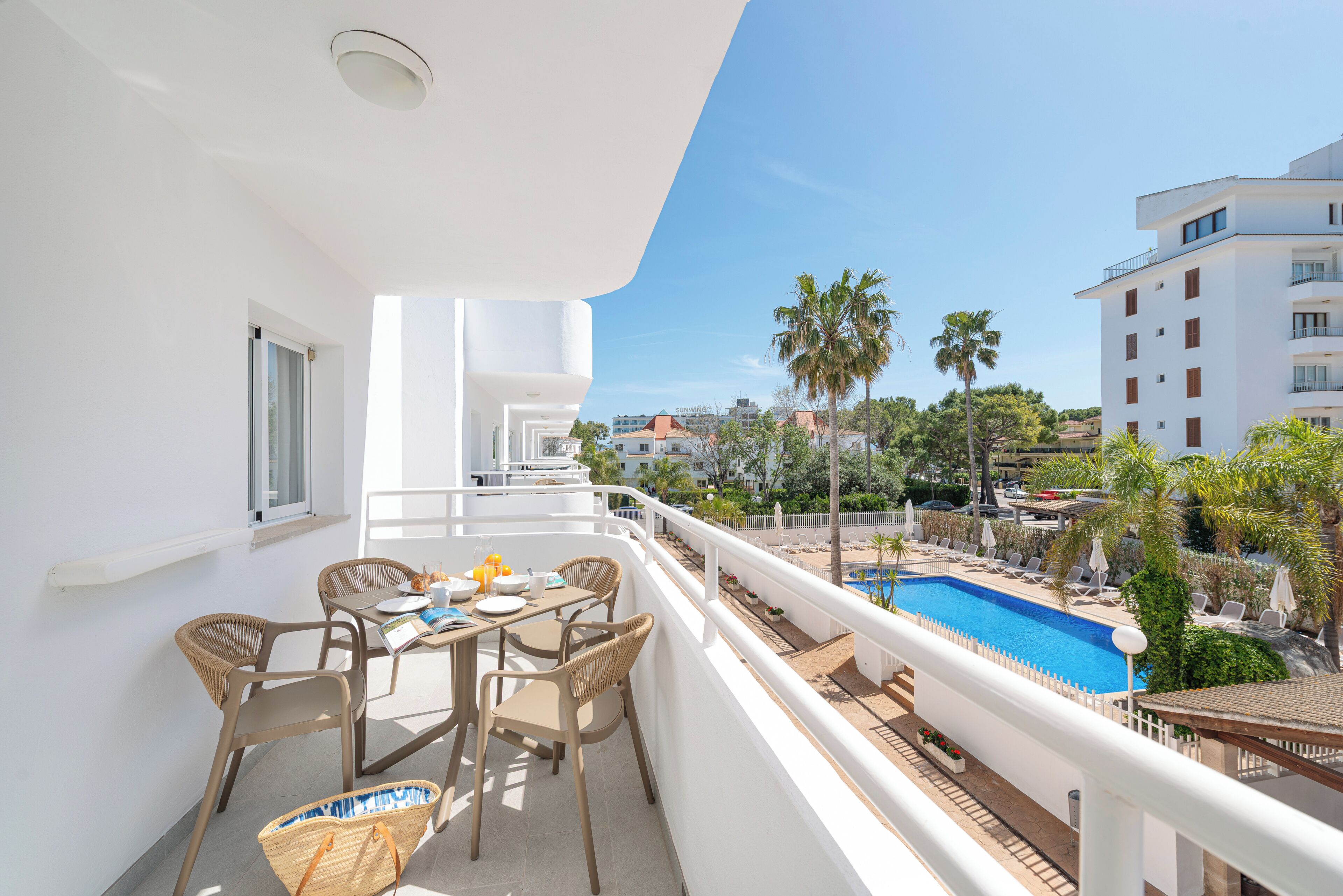 Un balcón con mesa y sillas, una piscina y vistas a palmeras y edificios.
