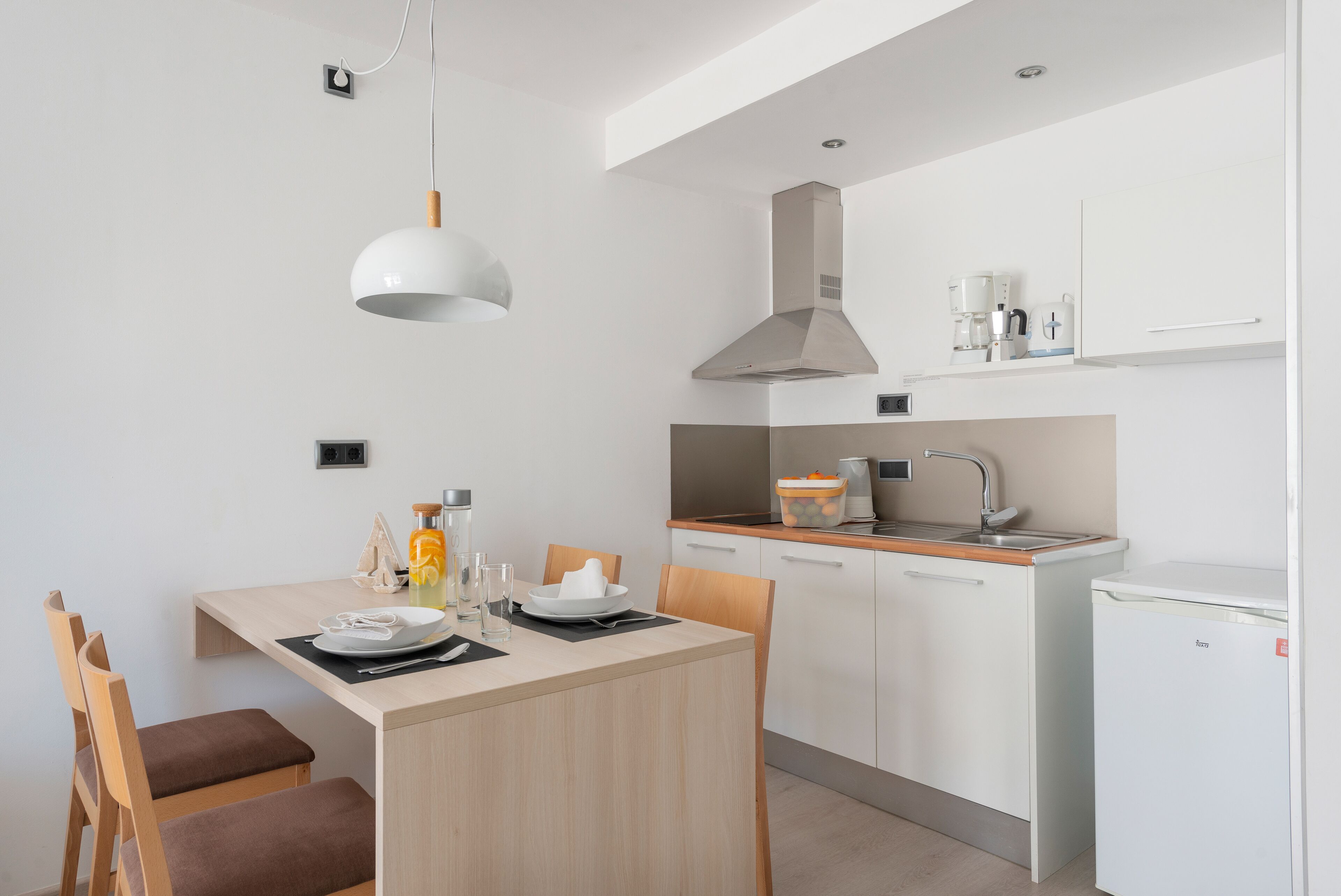 A modern kitchen with white cabinets, a built-in oven, and a sink. There is a dining table with chairs and a pendant light above it.