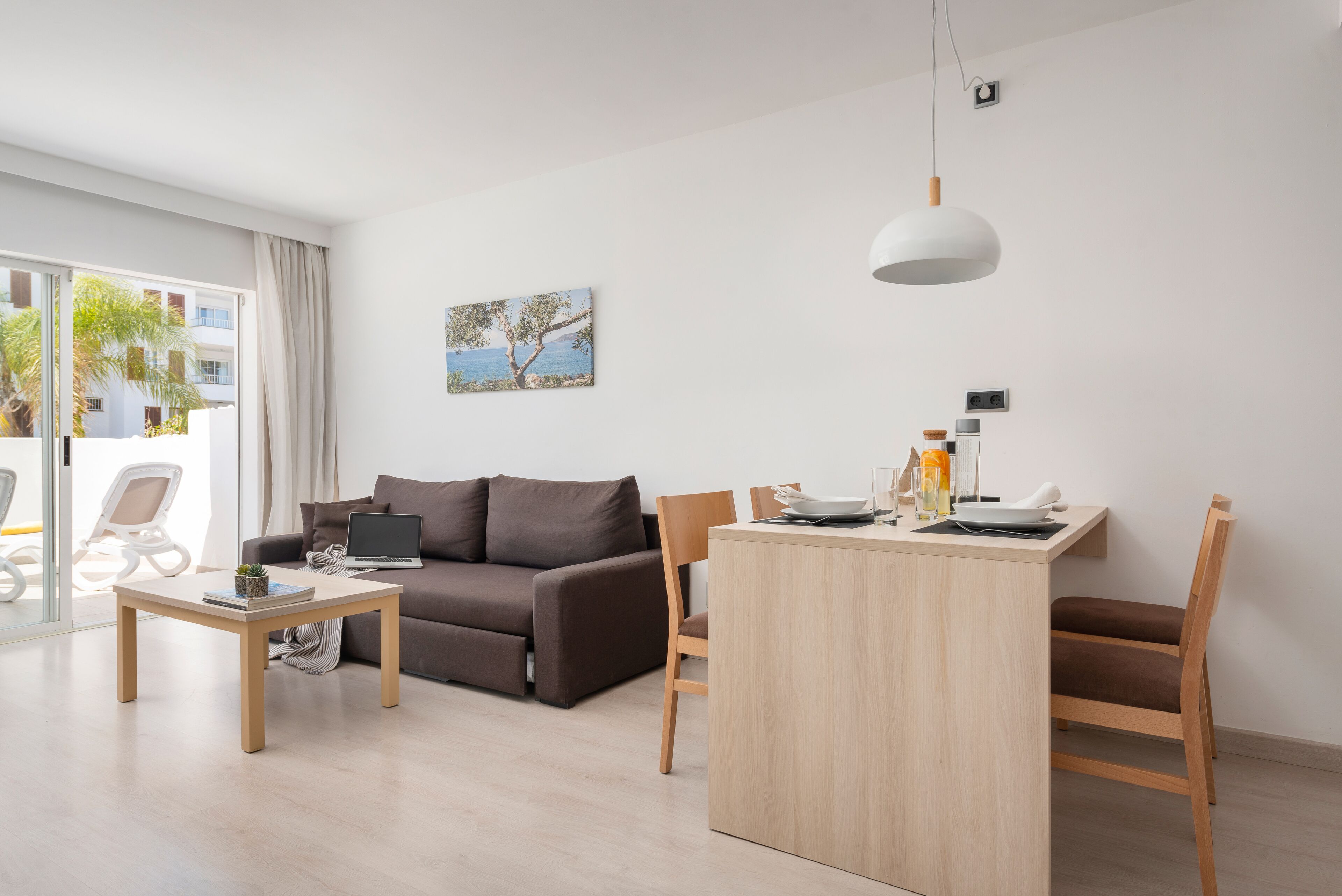 Un salon moderne avec un canapé, une table à manger et une chaise. On peut voir un ordinateur portable sur le canapé, une petite table avec une plante et un tableau au mur.
