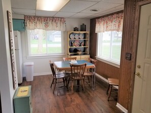 A dining area with a table and chairs, a trash bin, a shelf with decorative items, and a window with blinds. - Open concept 2 bedroom ranch with attached garage and deck (Essexville)