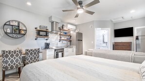 A modern bedroom with a large bed, a ceiling fan, and a kitchenette in the background.