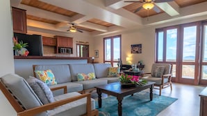 A spacious living room with a large sofa, two armchairs, a coffee table, and a ceiling fan. There is a kitchen area with wooden cabinets and a refrigerator. Large windows provide natural light and a view of the outdoors.
