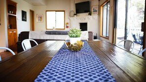 A dining table with a blue patterned runner, a white vase with a plant, and a wooden cabinet in a room with a fireplace and a television on the wall.