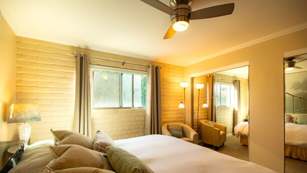 Hotelkamer met een groot bed, een plafondventilator, een raam met gordijnen, een stoel, een lamp en een spiegel.