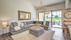 A modern living room with a sectional sofa, a round ottoman, a chandelier, and a large window with a view of a golf course. - Palm Desert Escape w/ Patio, Grill & Fire Pit (Palm Desert)