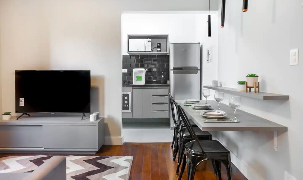 A modern kitchen with a built-in oven, microwave, and refrigerator. There is a dining area with a table set for two, and a TV on a stand.