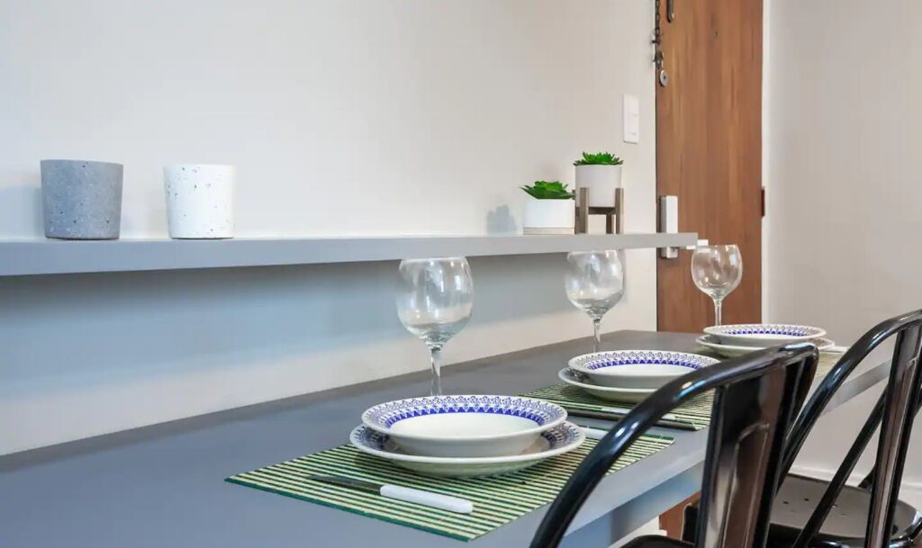 A dining table set with plates and wine glasses, a green placemat, and a small potted plant on a white shelf.