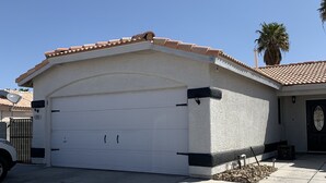 A single-story house with a white garage door, a tiled roof, and a small garden area with a palm tree. - Coming soon (North Las Vegas)