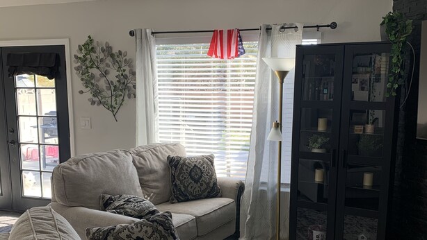 A living room with a beige sofa, patterned cushions, a glass cabinet with decorative items, and a view through the window. - Coming soon (North Las Vegas)