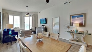 Ett modernt vardagsrum med ett matbord i trä, vita stolar, en blå soffa, en takfläkt, en tv och ett stort fönster med gardiner.