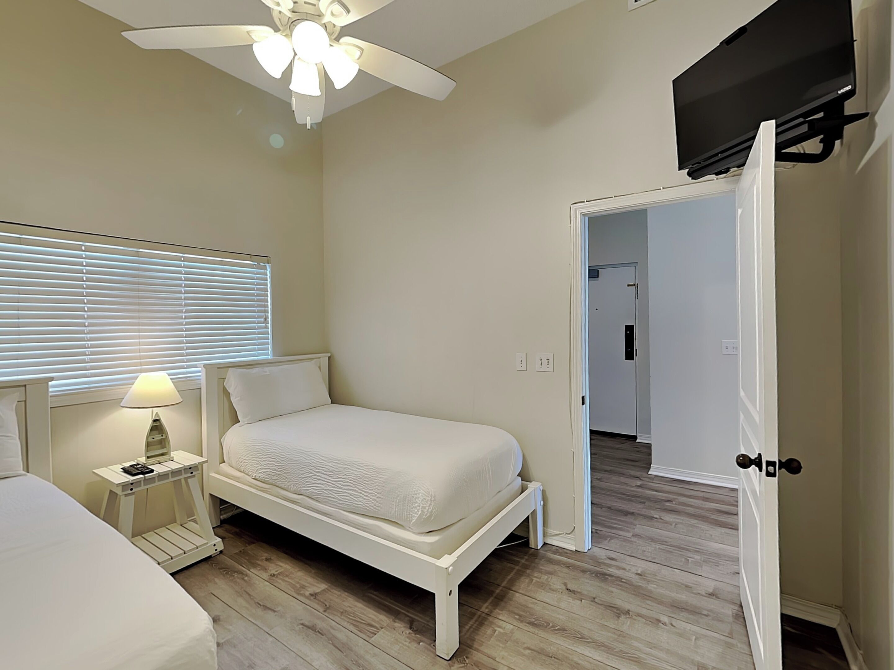 Un dormitorio con cama, mesita de noche, lámpara, ventilador de techo y una puerta abierta que da a otra habitación.