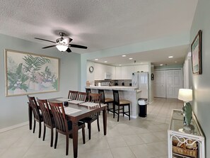 A dining area with a wooden table and chairs, a ceiling fan, and a kitchen in the background.