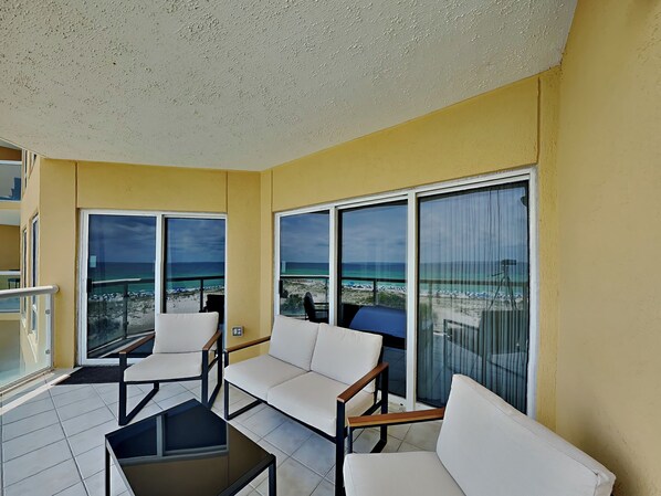A balcony with white cushioned chairs and a glass table, overlooking a beach with clear skies.