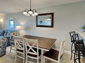 A dining area with a wooden table and white chairs, a hanging light fixture, and a framed mirror on the wall.