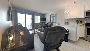En moderne stue med en sofa, et glasbord og et spiseområde med stole. Der er et køkken med hvide skabe og en lysekrone.