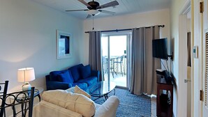 A living room with a blue sofa, a beige armchair, a glass coffee table, a TV on a stand, and a ceiling fan. - Baywatch B1 | Easy Access to the Sand, Community Pool! (Pensacola Beach)