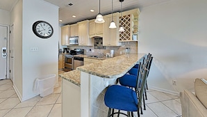 A modern kitchen with a central island, bar stools, and pendant lighting. - Baywatch B1 | Easy Access to the Sand, Community Pool! (Pensacola Beach)