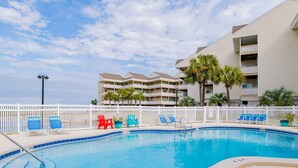A swimming pool with blue and red lounge chairs, a white fence, and a multi-story building with balconies and palm trees in the background. - Baywatch B1 | Easy Access to the Sand, Community Pool! (Pensacola Beach)