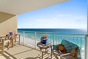 A balcony with a view of the ocean, a table with a cooler, and a lounge chair with a hat and a striped towel.