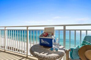 A balcony with a view of the ocean, a cooler with drinks, and a chair with a striped cushion. - Relax in Style | Gulf-View 1BR Condo at Emerald Beach | Summer Specials (Panama City Beach)