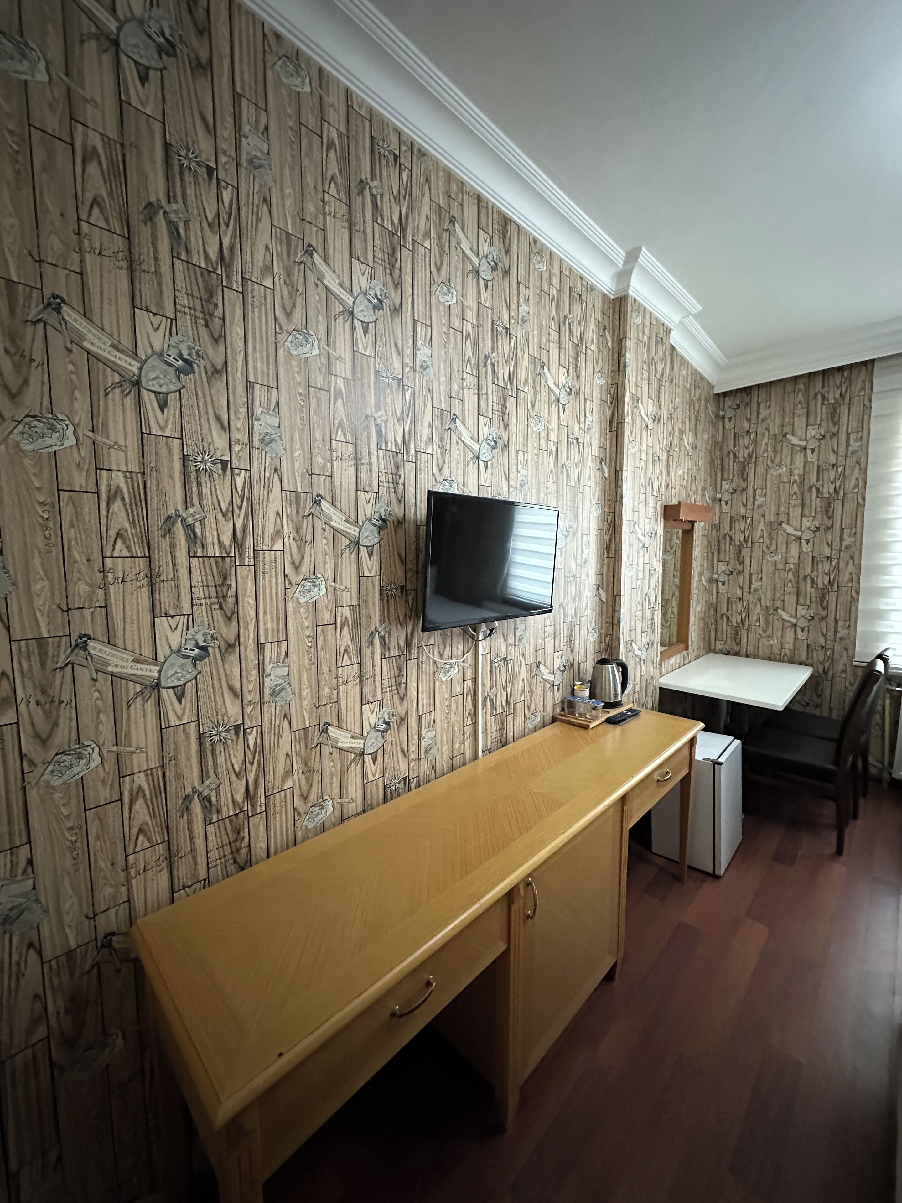 Una habitación compacta con pared de madera, un televisor de pantalla plana sobre un escritorio de madera, un refrigerador pequeño y una mesa blanca con una silla.