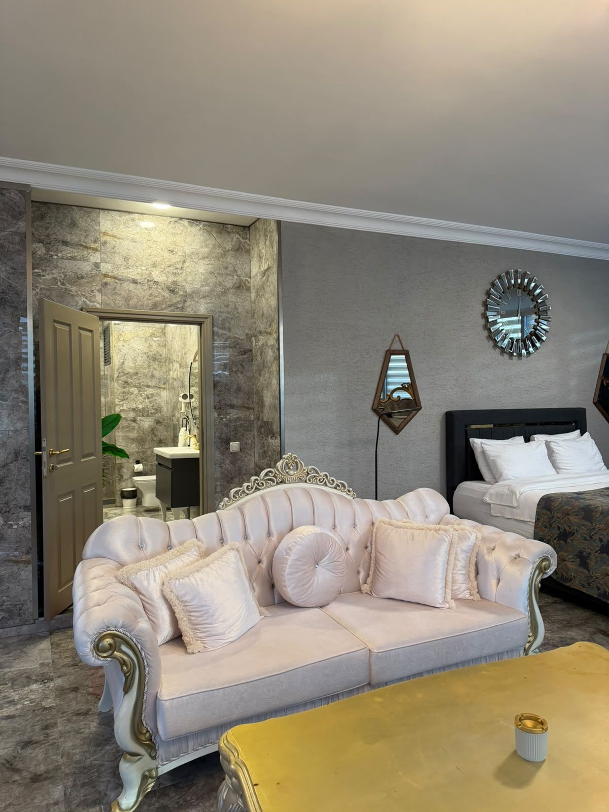 Une pièce avec un canapé beige, une table basse jaune, un lit, une horloge murale et une salle de bain visible à travers une porte ouverte.