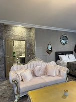 Una habitación con un sofá color beige, una mesa de centro amarilla, una cama, un reloj de pared y un baño visible a través de una puerta abierta.
