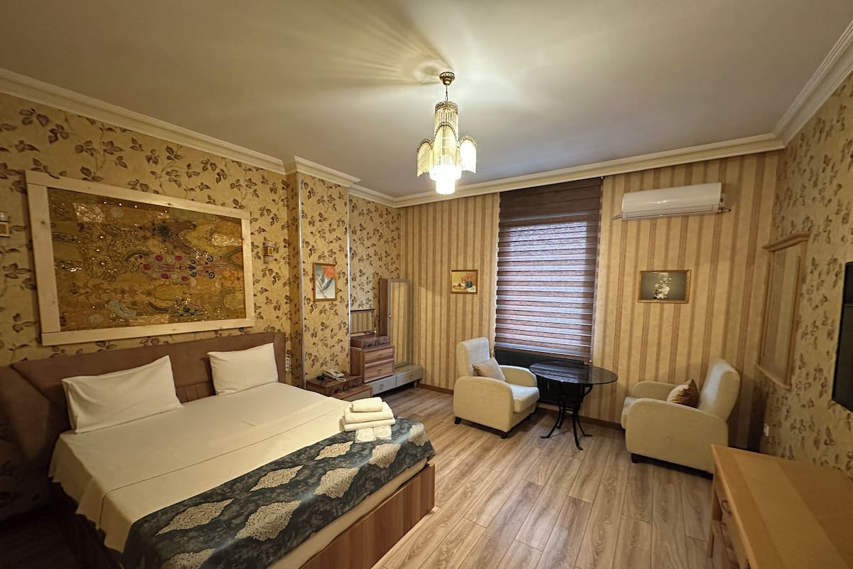 A hotel room with a large bed, two armchairs, a small table, a mirror, and a window with blinds.