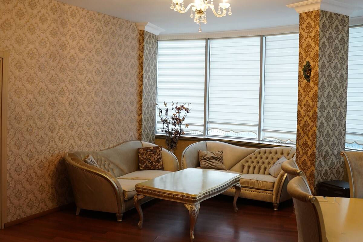 A room with a patterned wallpaper, a chandelier, a sofa, a dining table, and a window with blinds.