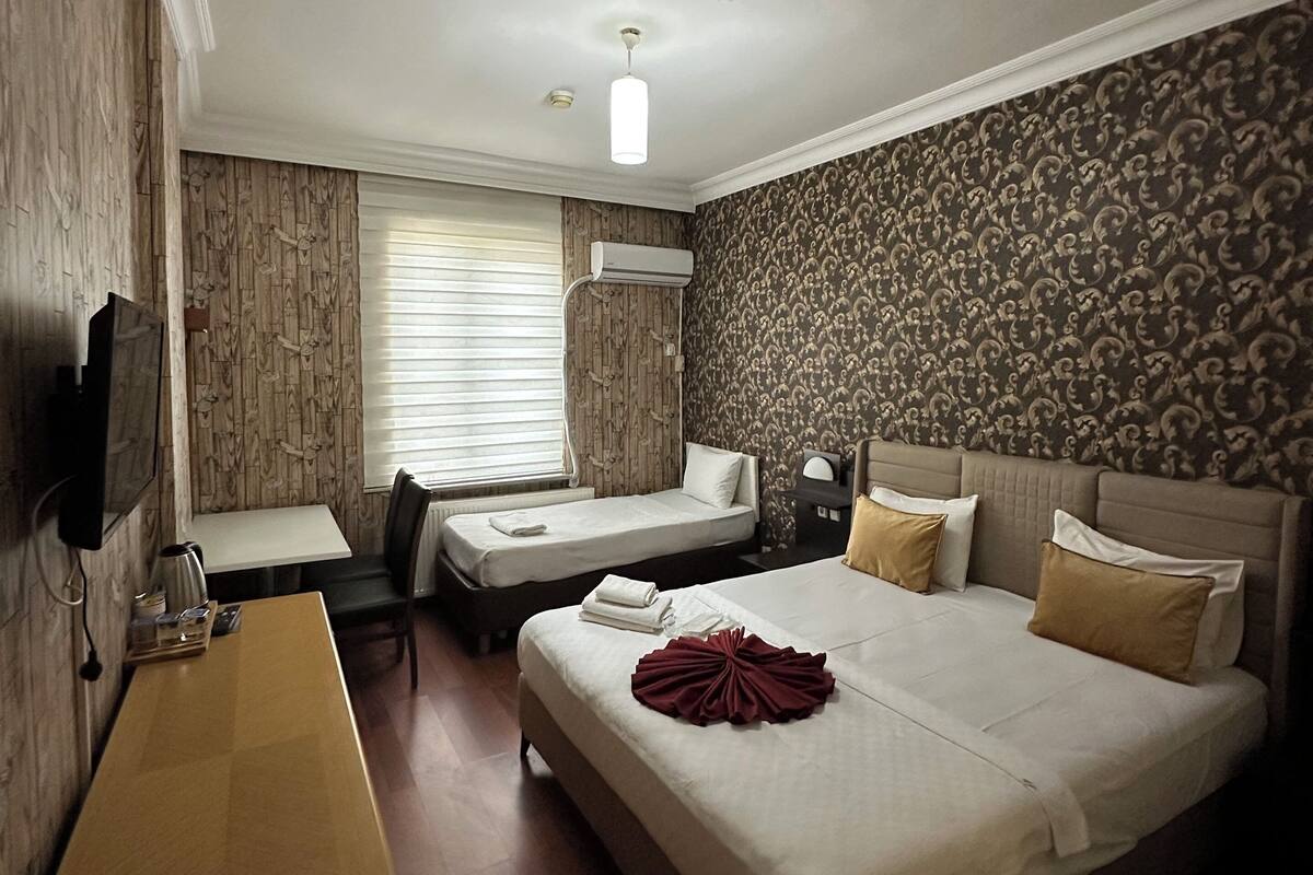 A hotel room with a bed, a desk, and a chair. There is a window with blinds, an air conditioning unit, and a television mounted on the wall.