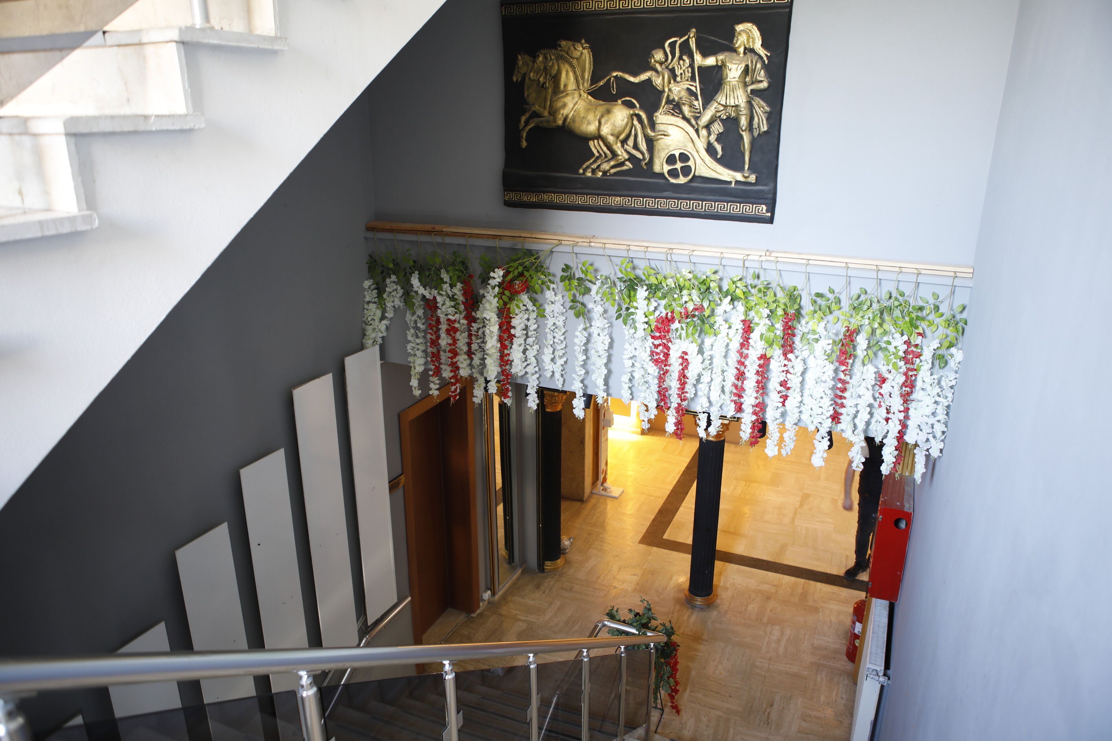 Una escalera con barandilla de metal, un estandarte decorativo con escena clásica y una guirnalda floral colgada arriba.