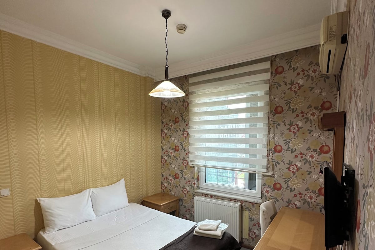 A bedroom with a bed, bedside tables, a desk, a chair, a window with blinds, and a radiator.