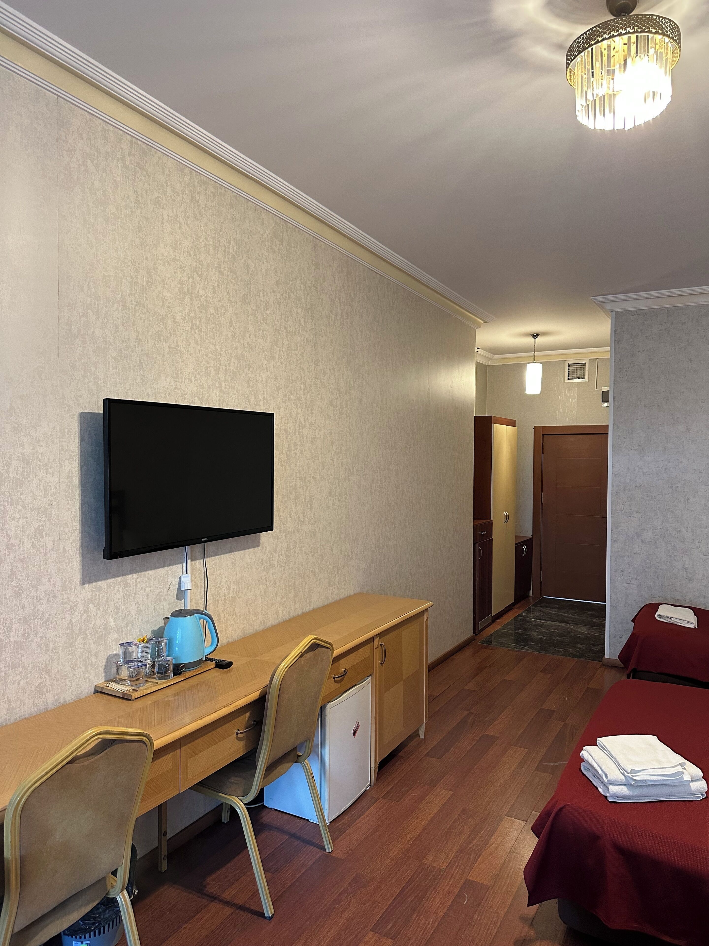 A hotel room with a wooden desk, a television, a small refrigerator, a chair, a bed with a red bedspread, and a hallway leading to another room.