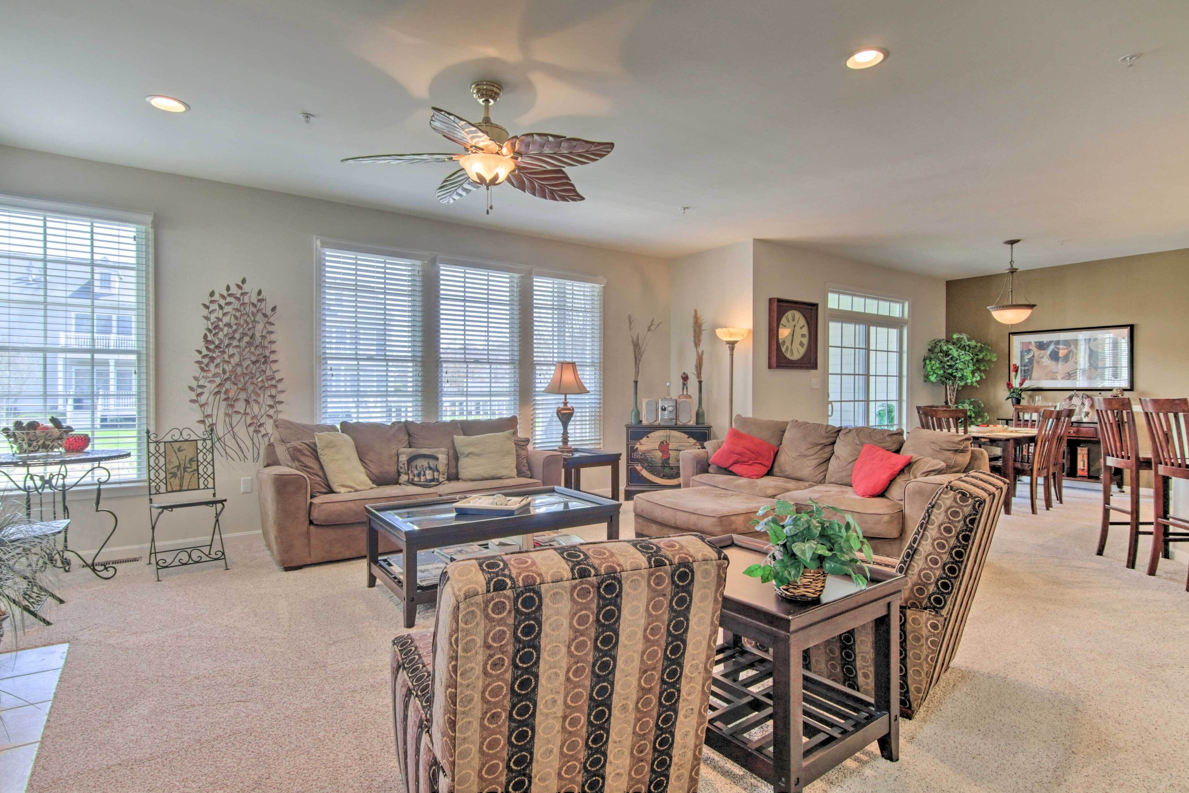 A spacious living room with a ceiling fan, large windows with blinds, a dining area with chairs and a table, and a coffee table.