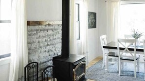 A rustic interior with exposed wooden beams, a black wood-burning stove, white walls, and a dining area with white chairs. - The White Barn (Julian)