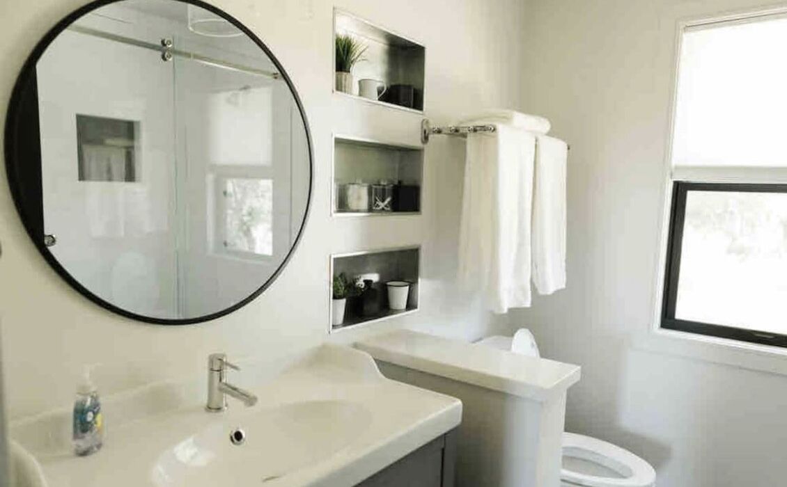 A bathroom with a round mirror, a sink, a toilet, and shelves with toiletries.