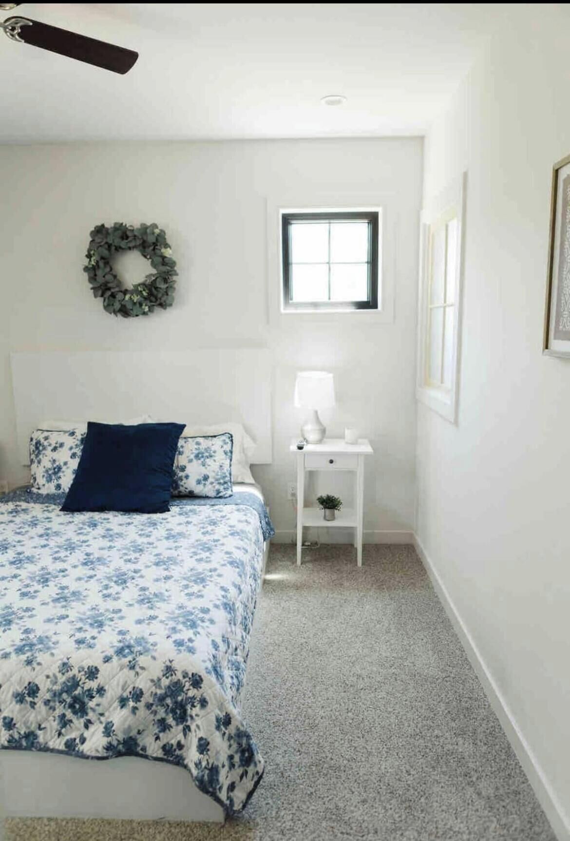 Une chambre à coucher avec un lit, une table de chevet, une fenêtre et une couronne décorative accrochée au mur.