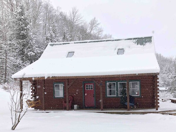 Una cabaña de madera con techo nevado, un porche delantero y una puerta roja.