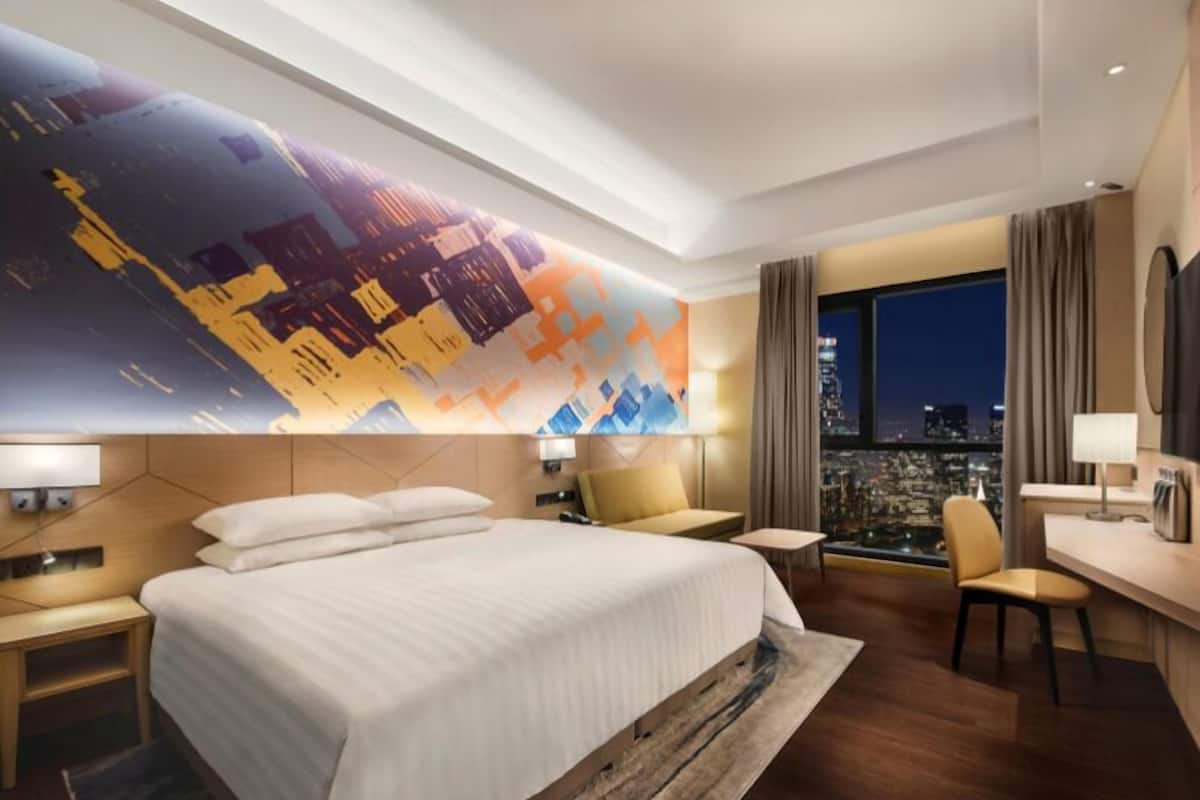 Habitación de hotel moderna con una cama grande, un escritorio y una silla. Se aprecia una vista panorámica de la ciudad a través de la ventana.