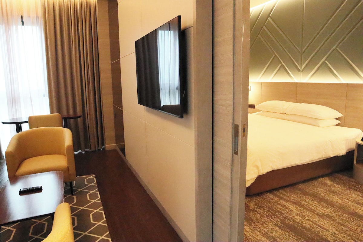 Una habitación de hotel con una cama, un televisor montado en la pared, una zona de estar con una silla y una mesa, y una alfombra estampada.